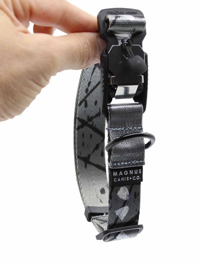 A silver nylon strap dog collar by MAGNUS Canis with black spatter print design and a magnetic buckle being held vertically.